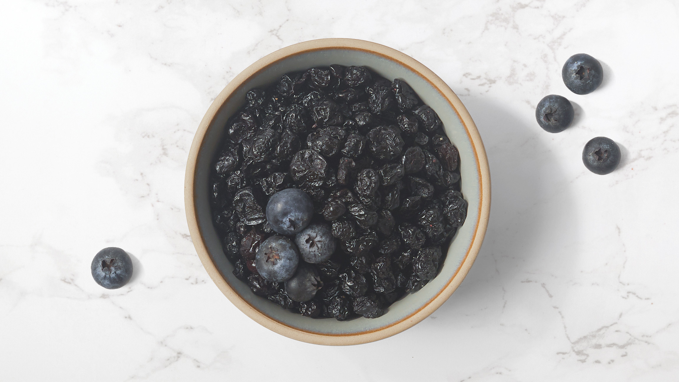 Dried blueberries in a ceramic bowl with fresh blueberries scattered on a marble counter