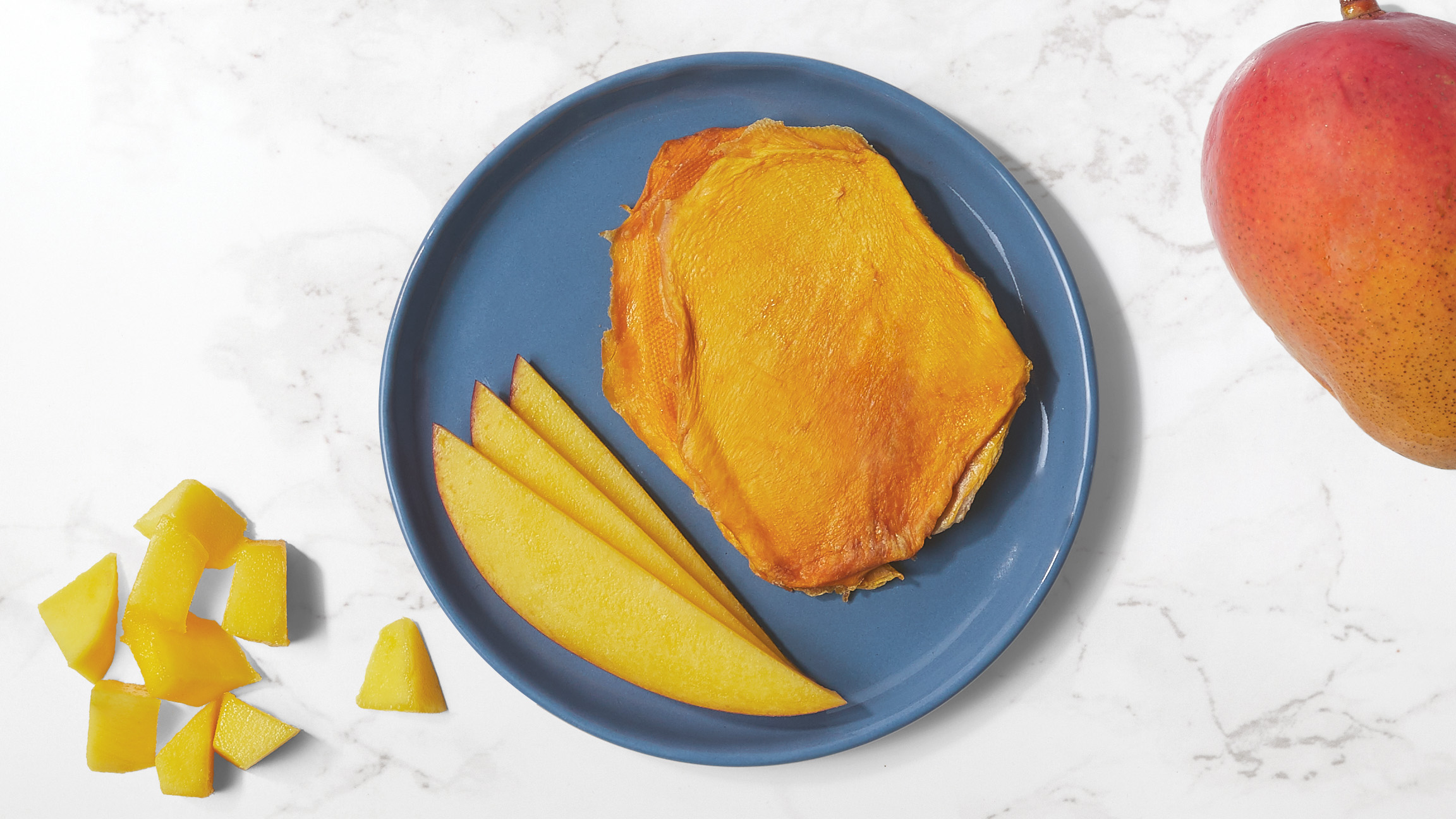 Dried and fresh mango slices on a blue plate with fresh pieces of mango and a whole mango on a white marble counter top