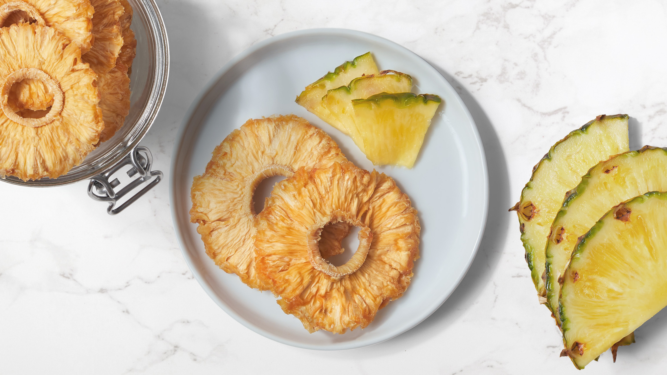 Dried pineapple rings and fresh pineapple slices on a white plate with fresh pieces of pineapple and a jar of dried pineapple rings on a white marble counter top