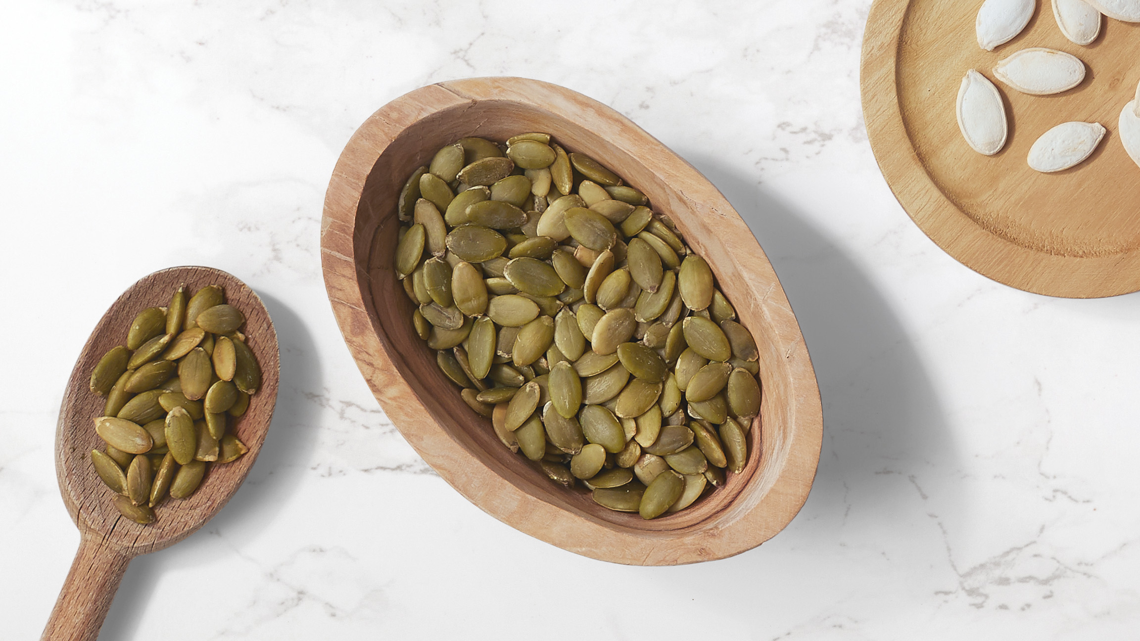 Pumpkin seeds in a wooden oval bowl accompanied with a wooden spoonful of pumpkin seeds on a white marble counter top