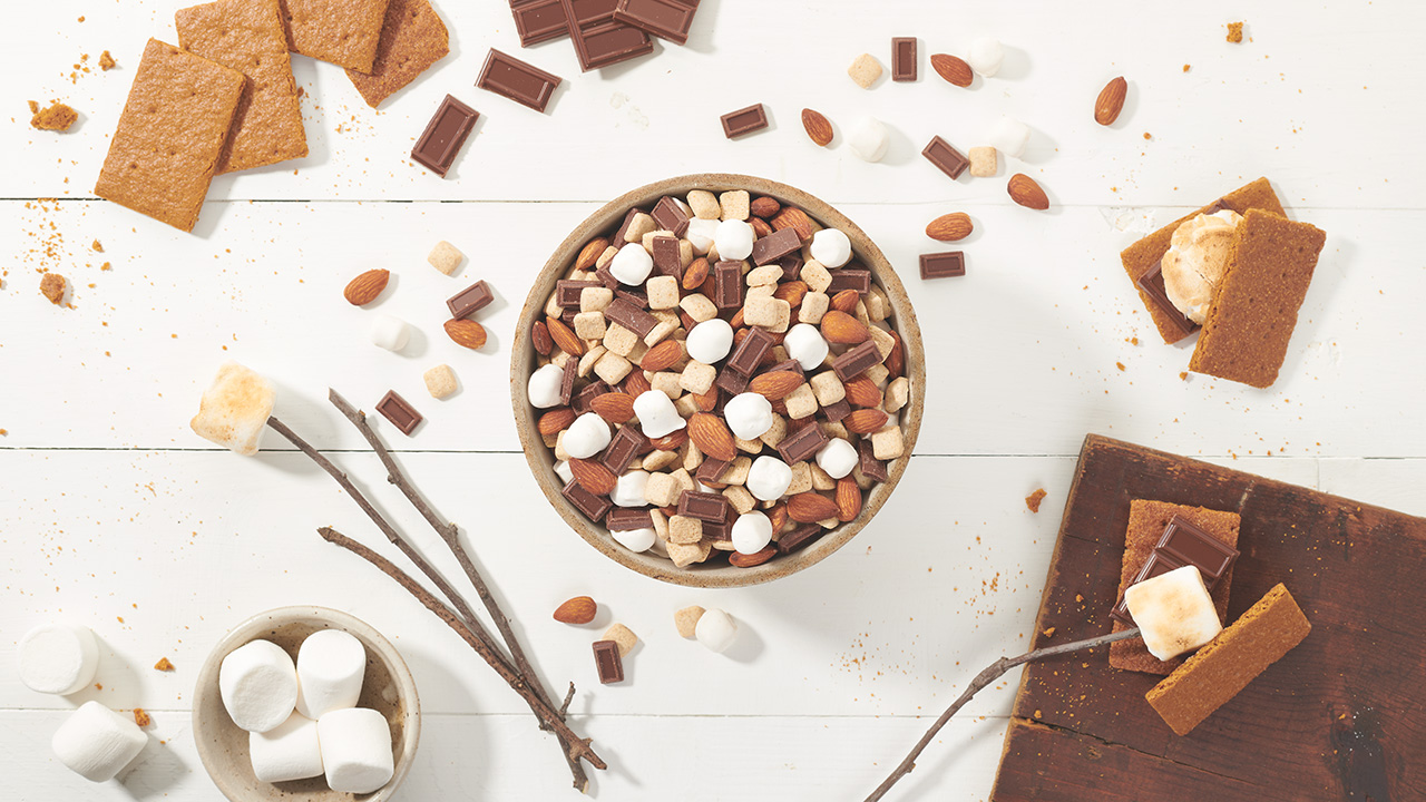 Bowl of S'more trail mix surrounded by scattered almonds, marshmallows, chocolate pieces and graham cracker pieces on a tabletop